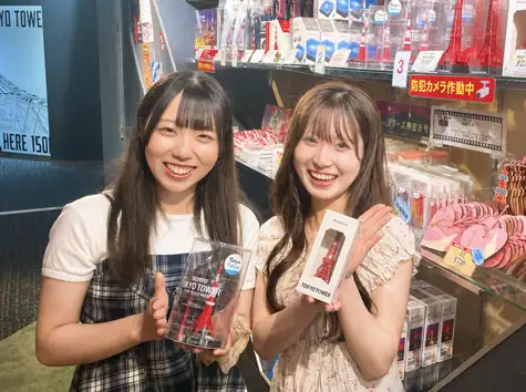 Two women smiling as they hold miniature Tokyo Tower models, with rows of amulets and souvenirs displayed behind them
