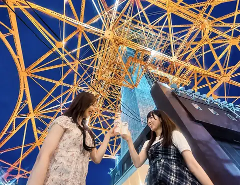 Two women drinking as they look up at the orange-lit steel frame of Tokyo Tower