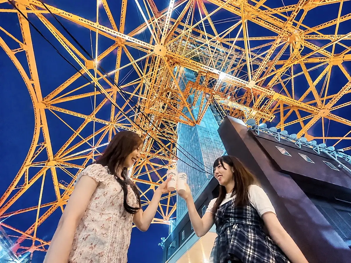 Two women drinking as they look up at the orange-lit steel frame of Tokyo Tower