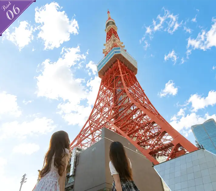 Two women seen from behind, looking up at the towering base of Tokyo Tower against a clear blue sky