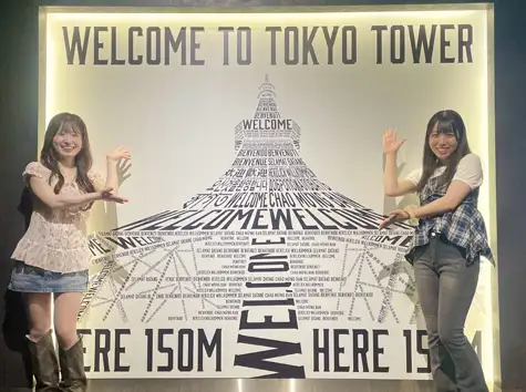 Two women smiling and taking photos together at the Tokyo Tower Main Deck photo spot, decorated in a tower shape with the words WELCOME TO TOKYO TOWER and welcome messages from around the world