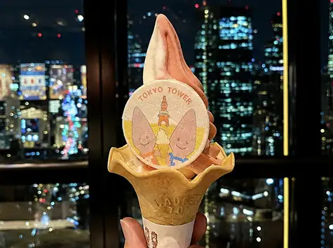 A premium Tokyo Tower-themed soft-serve ice cream with a coaster, held up against the Tokyo night view