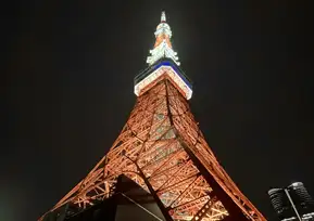 A dramatic low-angle photo of Tokyo Tower lit up against the sky. The tower is bathed in warm orange light, highlighting its intricate steel framework.