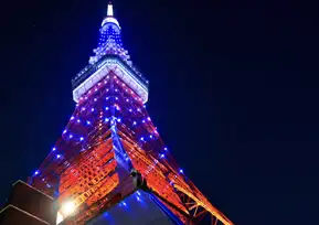 A photo looking straight up at the tower against a dark late-night sky, lit up in vivid red and blue LED lights. The red steel framework is beautifully accented with points of blue light.